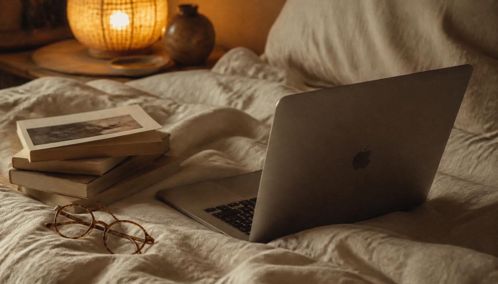 A laptop open on a linen bedspread in warm lamplight